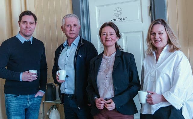 Fire av rådgiverne i Kon-text med kaffekopper foran døra med Kon-text-logoen.